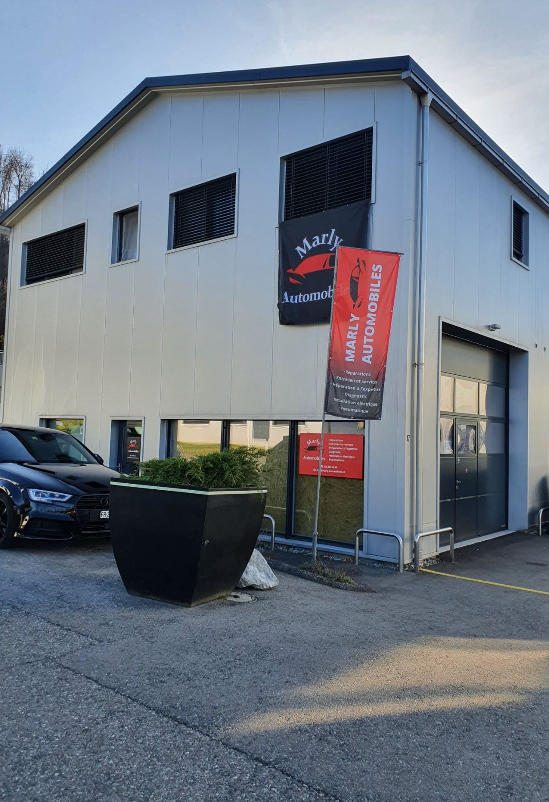 Façade du garage avec le drapeau Marly Automobiles et l'entrée de l'atelier